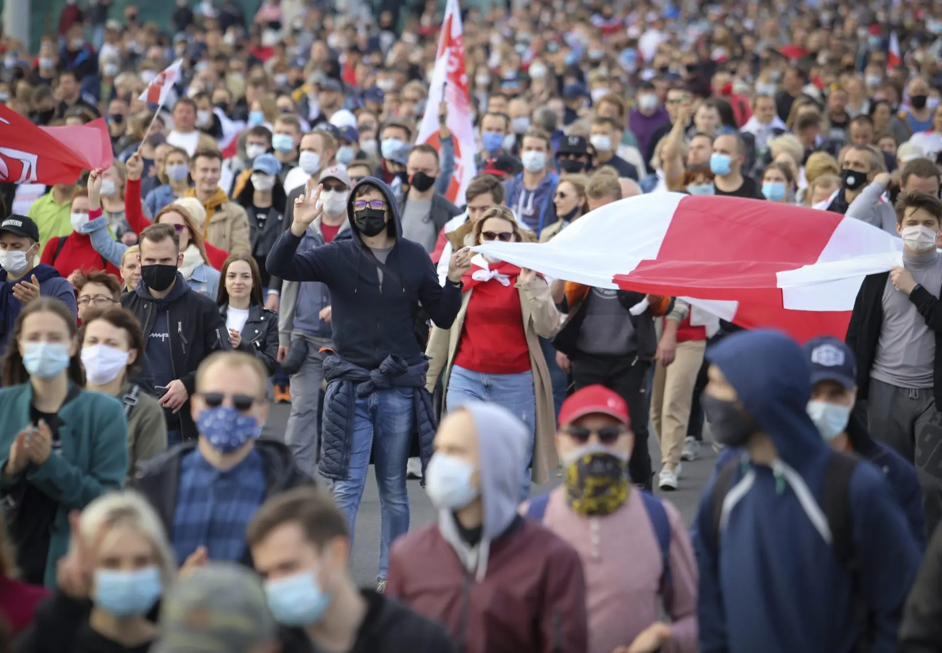 A large crowd of people, many wearing masks, march closely together holding red and white flags during a protest or demonstration. Some people are clapping and raising their hands.