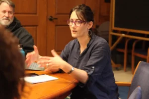 A woman with glasses and hoop earrings gestures while speaking about Harvard free speech at a meeting table, with a notepad and laptop in front of her. A man sits in the background, partially visible, against a wooden wall.