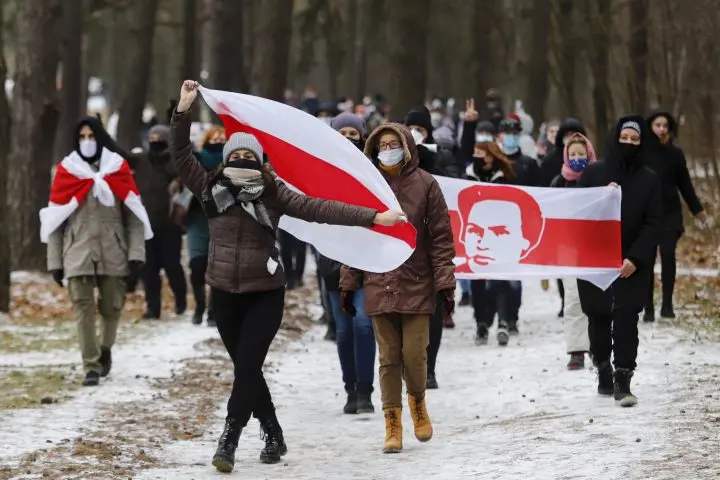 A group of people march in a snowy forest, many wearing masks. Some hold large red-and-white flags, one featuring a portrait of a man, suggesting a protest related to the Belarus congress or recent election.