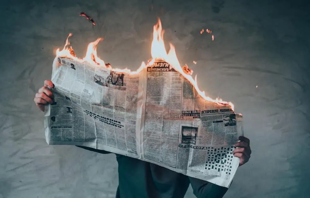 A person holds up an open newspaper that is burning along the top edge, with flames and smoke visible, against a plain gray background. The person’s face is hidden behind the newspaper.
