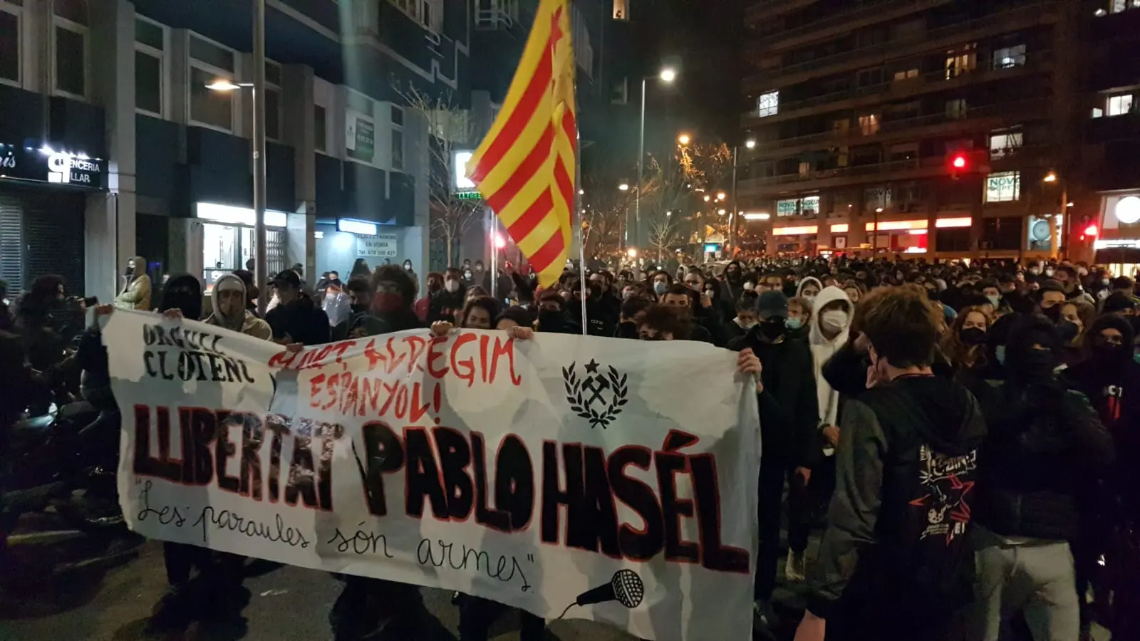 A large crowd protests at night on a city street, holding a banner reading Llibertat Pablo Hasél and a Catalan flag. Many people wear masks, and buildings with lights are visible in the background.