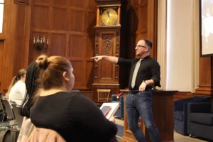 A man in a black shirt and tie stands and points while speaking to an audience in a wood-paneled room with a large clock during a Harvard free speech event. A woman in the foreground listens and takes notes on a tablet.