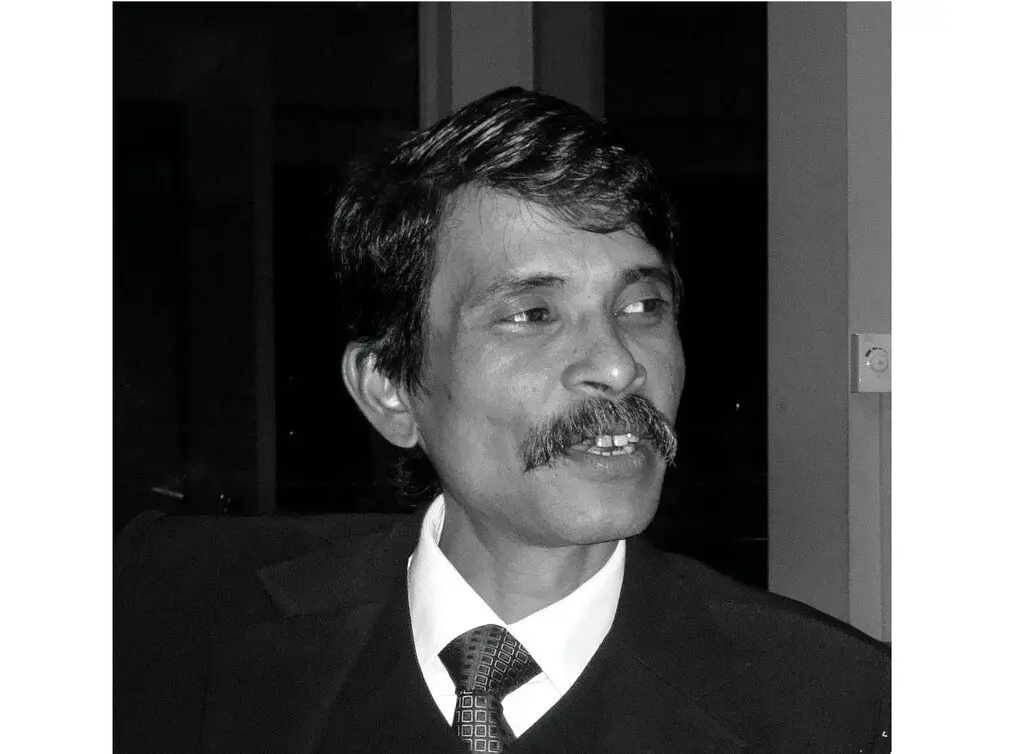 A man with dark hair and a mustache, dressed in a suit and tie, is looking slightly to his left. The photo is in black and white, and he appears to be mid-conversation or smiling subtly.