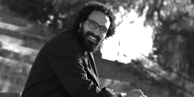 A man with curly hair, a beard, and glasses is smiling while sitting outdoors on steps. The image is in black and white.