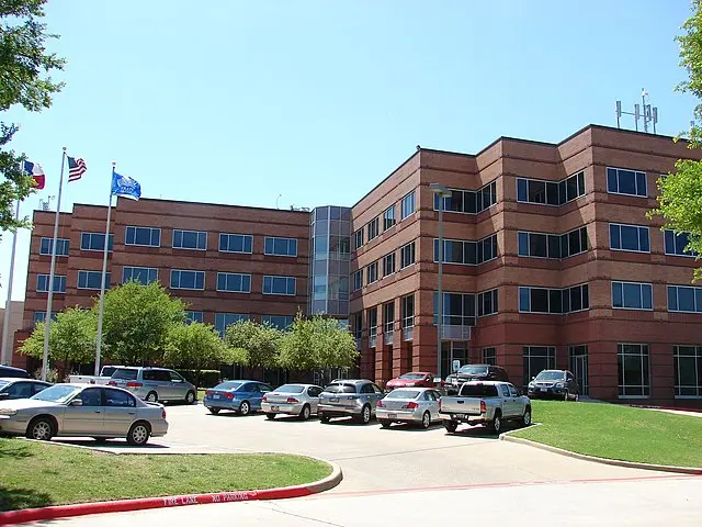 A large, modern brick office building with three stories and many windows, surrounded by trees. Several cars are parked in the lot, and three flags are flying near the entrance under a clear blue sky.