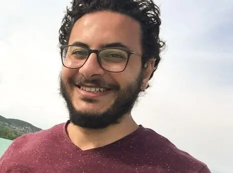 A man with curly dark hair, glasses, and a beard is smiling outdoors. He is wearing a maroon t-shirt, and the background shows a bright sky and a hint of landscape.