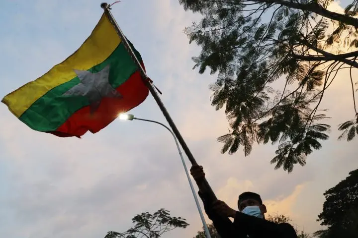 A person wearing a mask holds up the Myanmar national flag on a pole, silhouetted against a dusky sky with trees and a street light in the background.