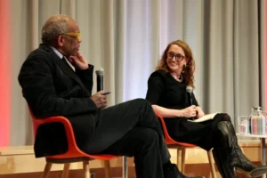 Two people sit in orange chairs on stage, each holding a microphone and engaged in a discussion about Harvard free speech. The man on the left wears glasses and a suit; the woman on the right, in glasses, black dress, and boots, smiles at him.