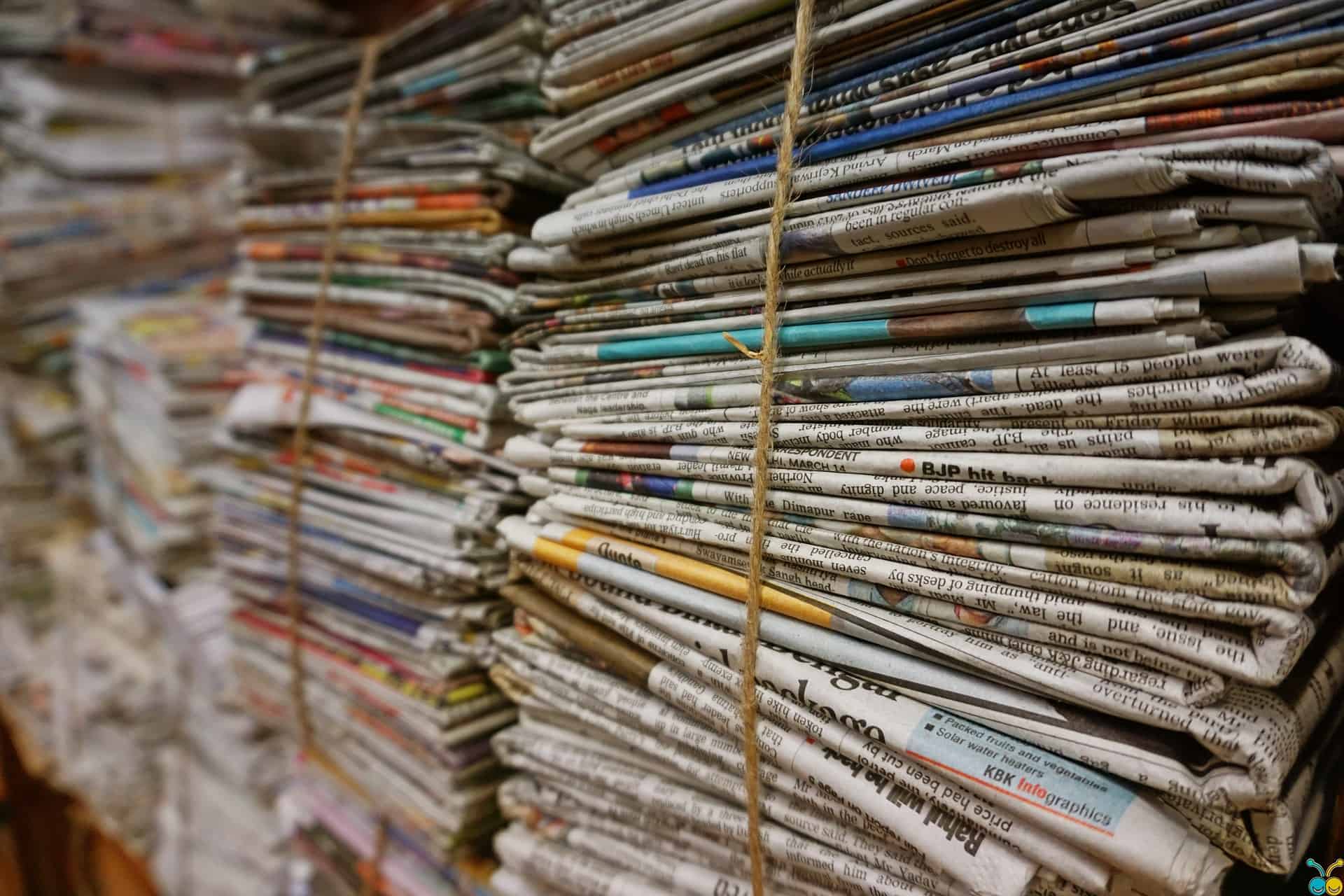 Stacks of old newspapers tied together with thick string, arranged in rows. The newspapers are neatly bundled, showing various headlines, articles, and colorful ads on the folded pages.