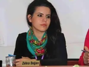 A woman with long dark hair, wearing a colorful scarf and black top, sits at a table with a nameplate that reads Zehra Doğan. She appears to be listening attentively.