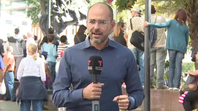 A male reporter in glasses and a blue shirt speaks into a microphone outdoors, with a crowd of people and trees visible in the background.