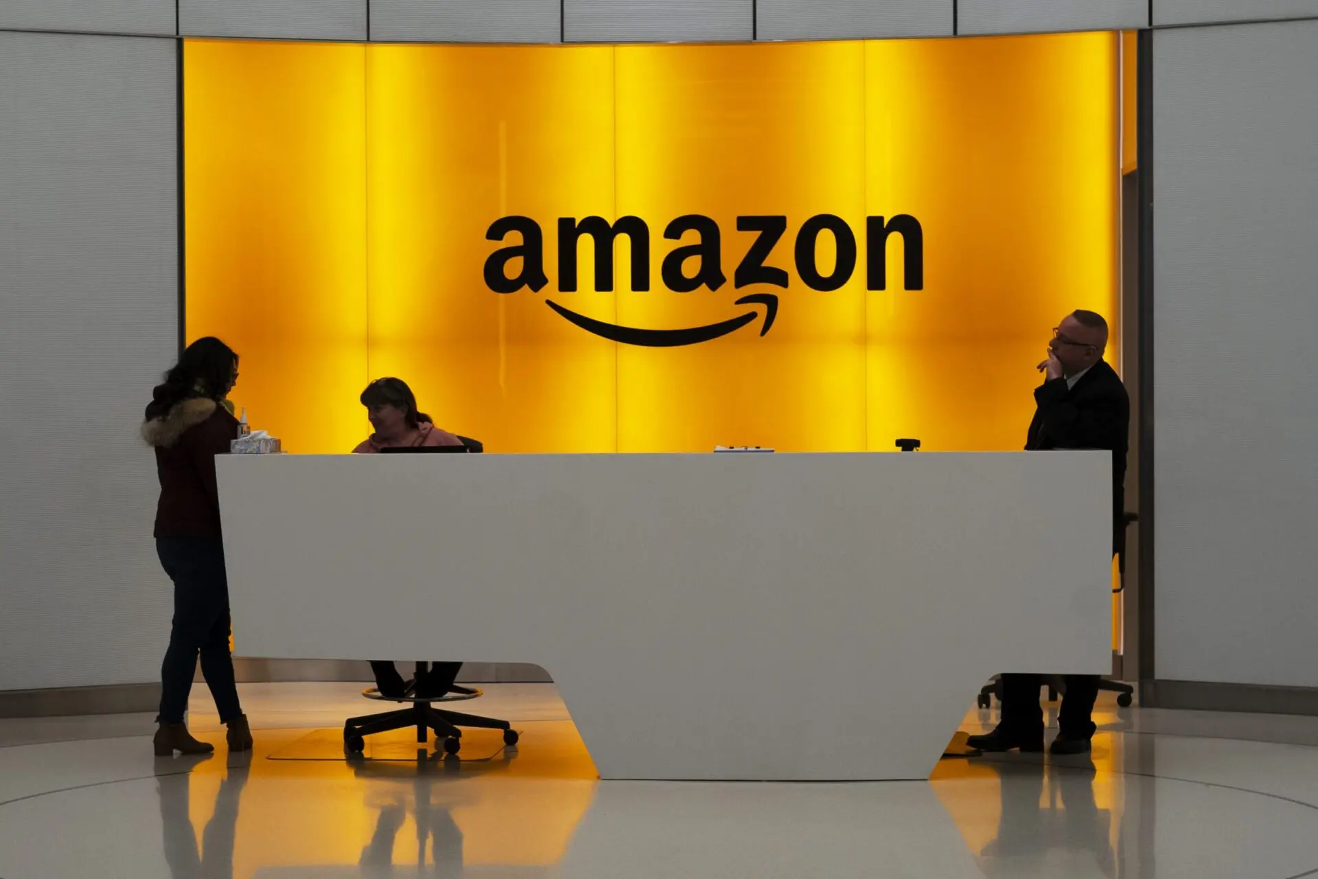 Three people stand and sit at a modern white reception desk in front of a large illuminated yellow wall with the black Amazon logo in the background.