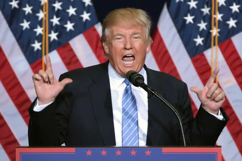 A man in a suit and striped tie speaks passionately at a podium with microphones, gesturing with both hands raised. Several large American flags are displayed in the background.
