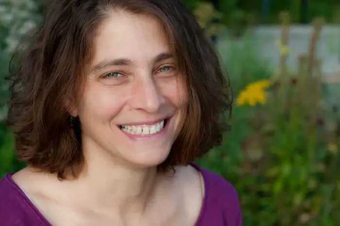 A woman with shoulder-length brown hair and green eyes smiles warmly at the camera, wearing a purple top. The background is blurred with greenery and yellow flowers.