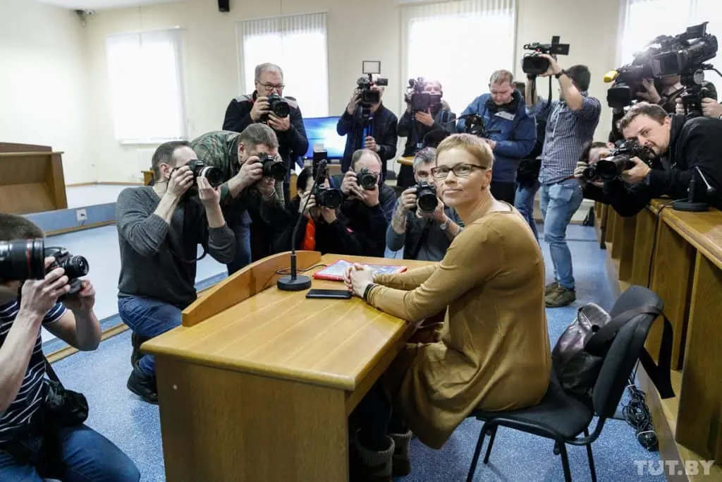 A woman sits at a courtroom desk, looking towards the camera, while several photographers surround her, taking pictures. The room is brightly lit with large windows in the background.