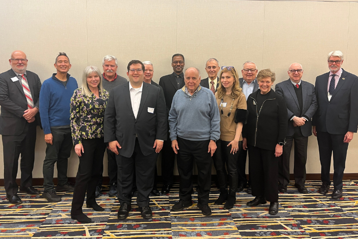A group of thirteen adults, dressed in business and business-casual attire, stand together and smile for a photo in front of a beige wall on a patterned carpet, following a discussion on university censorship.