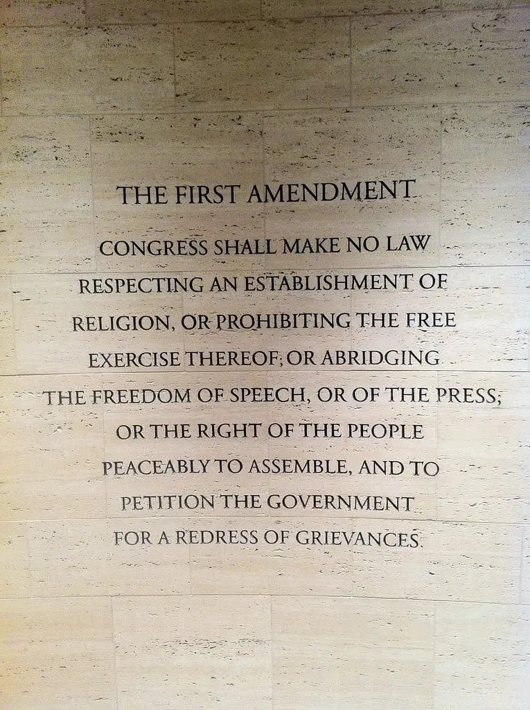 Text of the First Amendment inscribed on a beige stone wall, stating rights to religion, speech, press, peaceful assembly, and petitioning the government for grievances.
