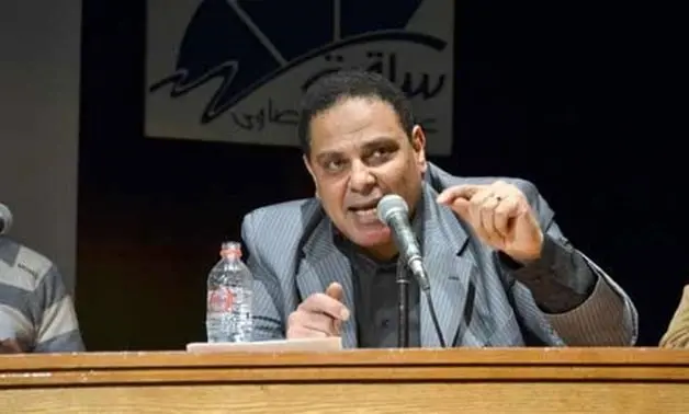 A man in a striped suit speaks passionately into a microphone at a panel table, gesturing with his hand; a water bottle and papers are in front of him, with a logo on the wall in the background.