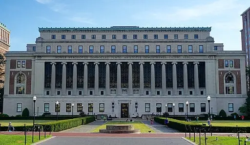 A large, neoclassical university library building with tall columns and many windows, surrounded by a green lawn and pathways. People are walking and sitting outside on a sunny day.