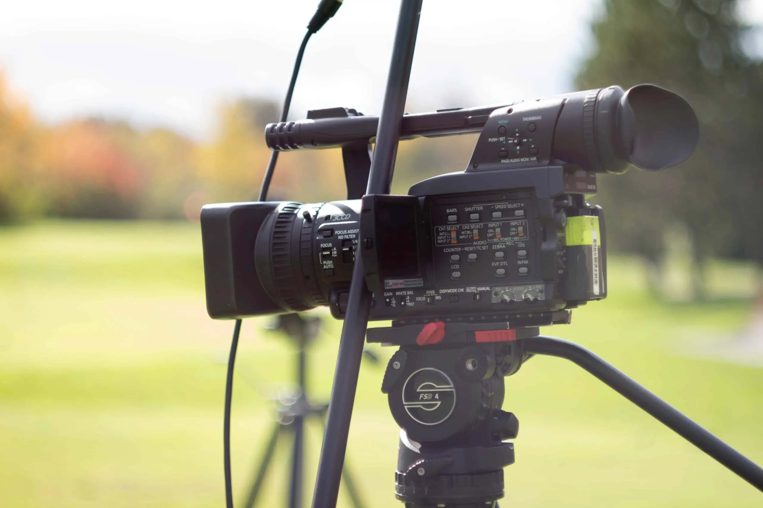 A professional video camera mounted on a tripod is set up outdoors on a grassy field, with trees blurred in the background and a microphone attached to the camera.