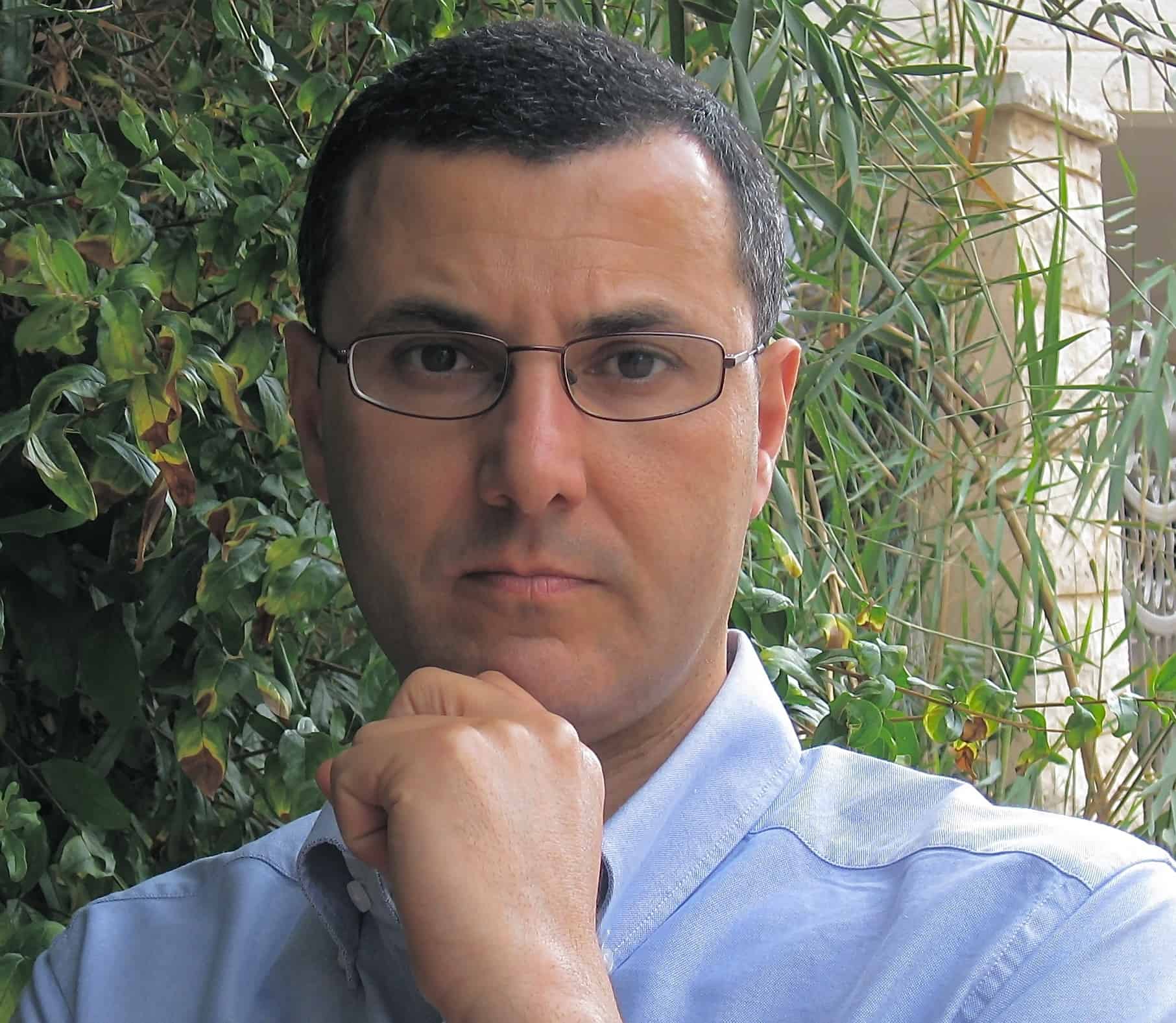 A person with short dark hair and glasses poses outdoors, wearing a light blue shirt, with one hand resting under their chin and green plants in the background.