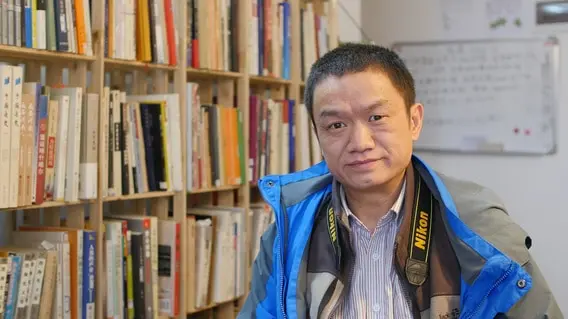 A man in a blue jacket with a Nikon camera strap sits in front of bookshelves filled with books, looking at the camera and smiling slightly. A whiteboard is visible in the background.