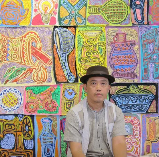 A man wearing a hat and a striped shirt sits in front of a vibrant wall covered with colorful, abstract paintings featuring various objects and patterns.