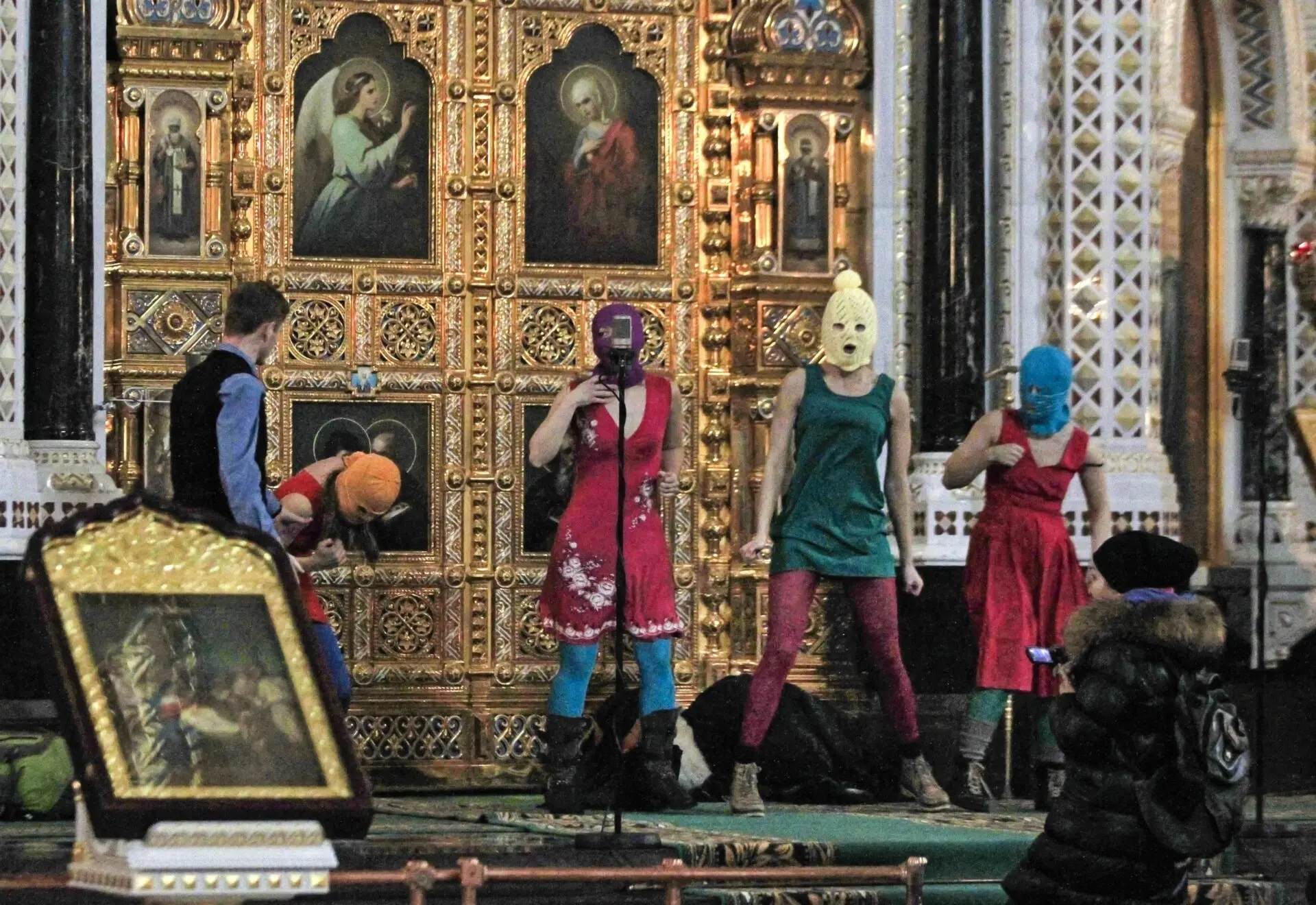 Five individuals in colorful dresses and balaclavas perform in front of an ornate religious iconostasis inside a church, while another person in dark clothing stands to the right taking a photo.