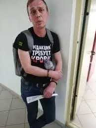 A person with a backpack leans against a white wall in a hallway, holding a rolled-up document. They are wearing a black T-shirt with white and red Cyrillic text.