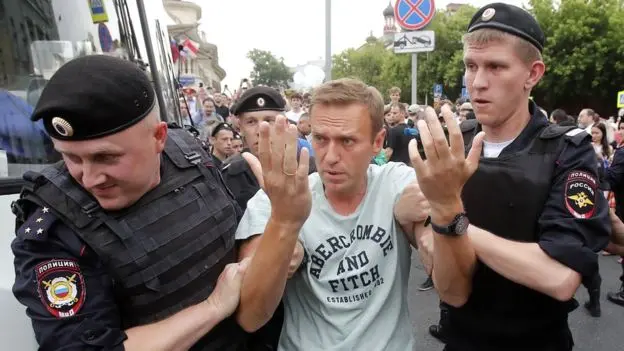 A man in an Abercrombie & Fitch shirt is being detained by two uniformed police officers in a crowded outdoor setting, with onlookers and buildings visible in the background.