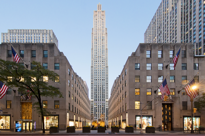A symmetrical view of the Rockefeller Center in New York City, with flags lining both sides and the tall central skyscraper rising in the background amid other office buildings at dusk.