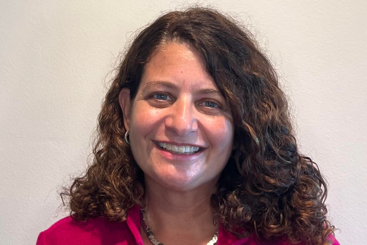 A woman with curly brown hair and a pink top smiles at the camera in front of a plain light-colored wall.