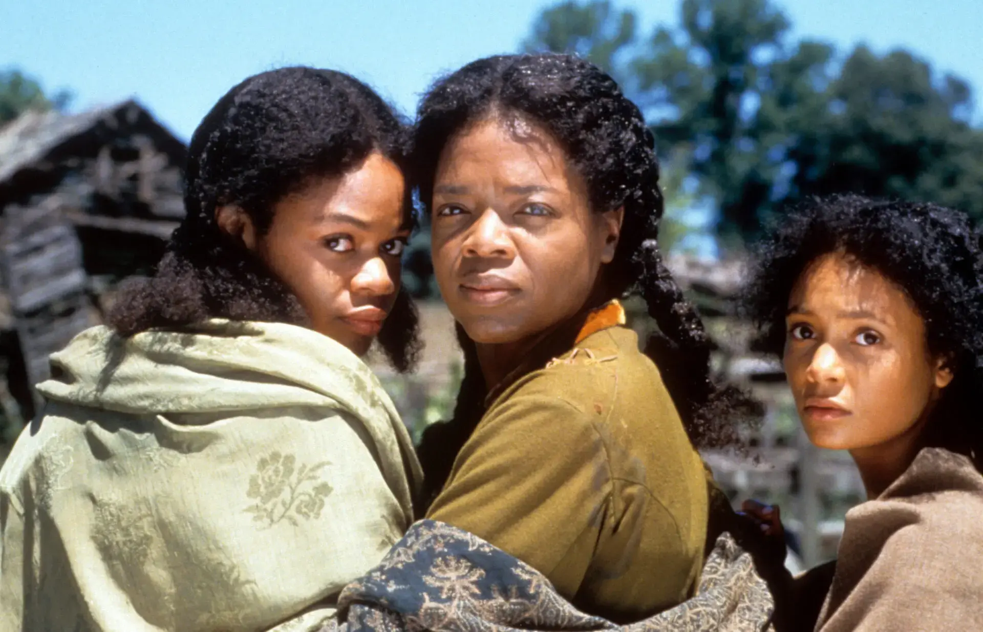 Three Black women with serious expressions stand closely together outdoors, wrapped in shawls, resembling a scene from banned book movies, with rustic wooden buildings and trees in the background on a sunny day.