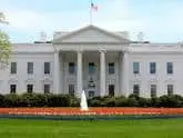The White House, a large white neoclassical building with columns, stands behind a lawn and a flower bed. An American flag flies on the roof against a blue sky.