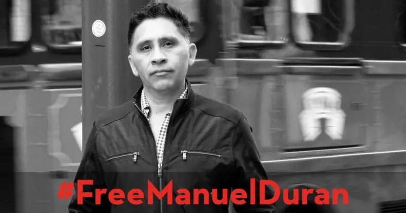 A man stands in front of a blurred background, wearing a zipped jacket and checkered shirt. Red text at the bottom reads #FreeManuelDuran. The image is in black and white.