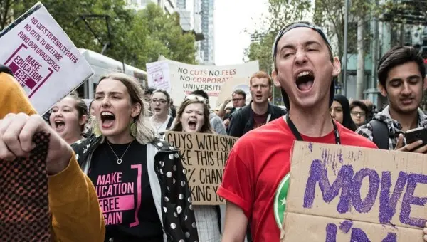 A group of young protesters march outdoors, holding signs and shouting. The signs call for free education and no cuts to university funding. The crowd appears passionate and determined, with trees and buildings in the background.