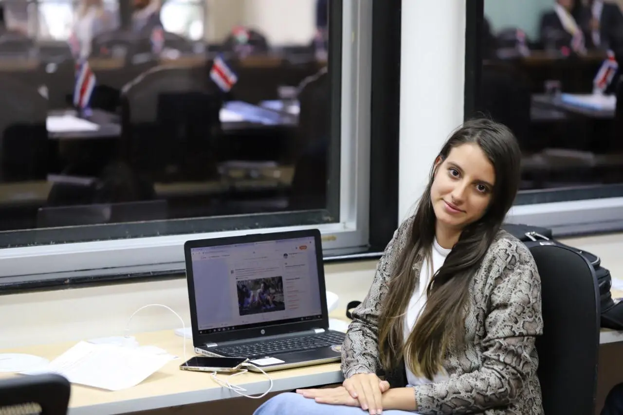 A woman with long brown hair sits at a desk, looking at the camera, with an open laptop in front of her. Behind her, there is a window showing a room with desks and small Costa Rican flags.