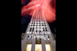 A tall building at night with red light streaks above it. Projected text on the building reads: “In a world where independent voices are increasingly stifled, pen is not a luxury. It’s a necessity.”.