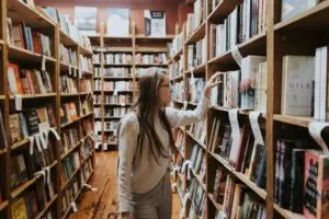 A woman with long hair and glasses stands in a bookstore aisle, reaching for a book on a wooden shelf filled with various books.