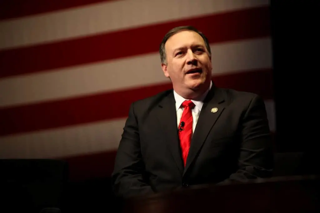 A man in a dark suit and red tie sits in front of a large, blurred American flag backdrop, looking slightly to the side.