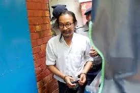 A man with long hair and glasses, wearing a white shirt and handcuffs, is escorted by police officers through a narrow passage with a brick wall.