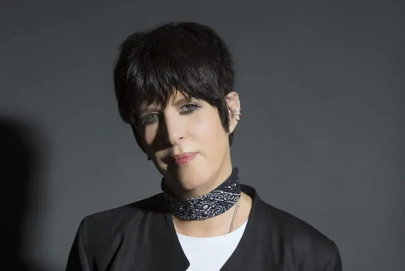 A person with short dark hair wearing a black jacket, a white shirt, and a patterned scarf around their neck stands against a plain gray background.