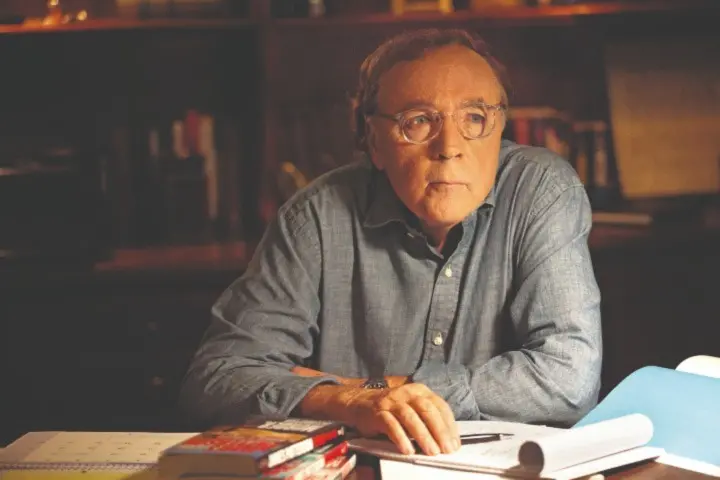 A man wearing glasses and a button-up shirt sits at a desk with books, papers, and a pen, looking thoughtful. Shelves filled with books are in the background.