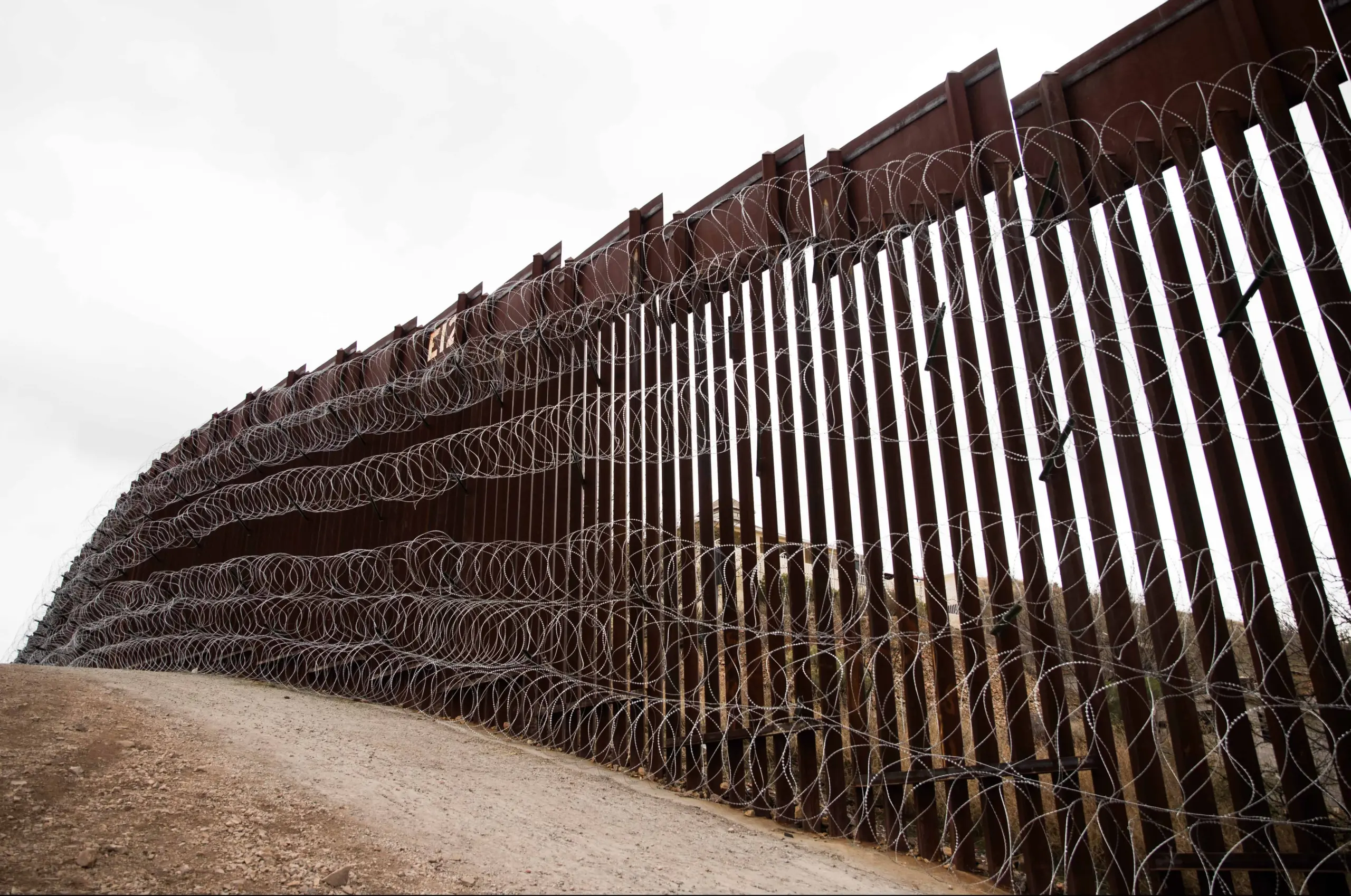 A tall metal border fence topped and lined with multiple layers of barbed and razor wire stretches across a dirt landscape under an overcast sky.