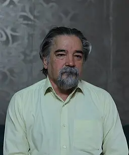 A middle-aged man with gray hair and a beard, wearing a light yellow button-up shirt, sits in front of a patterned gray wall, looking slightly serious.