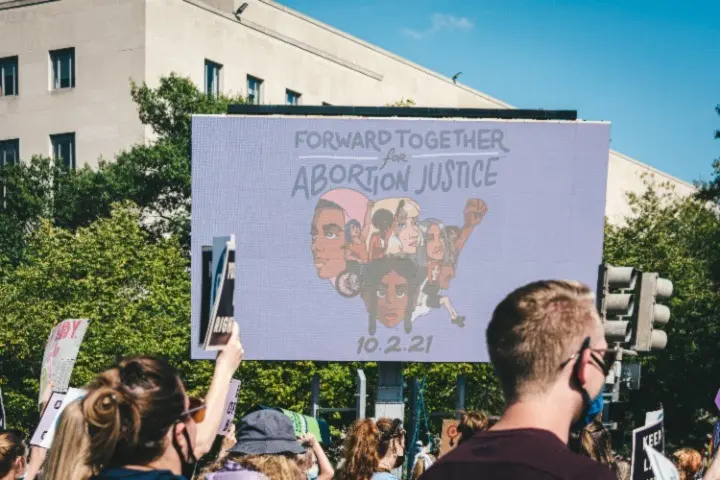 A crowd at a protest holds signs in front of a large screen displaying illustrated faces and the words Forward Together for Abortion Justice 10.2.21 with trees and a building in the background.