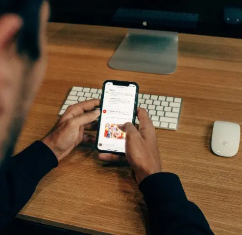 A person holds a smartphone and scrolls through social media while sitting at a wooden desk with a keyboard, mouse, and computer monitor in the background.
