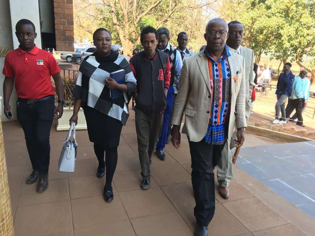 A group of people, including men and a woman carrying a white handbag, walk together outdoors on a paved walkway near a building, with trees and other people in the background.