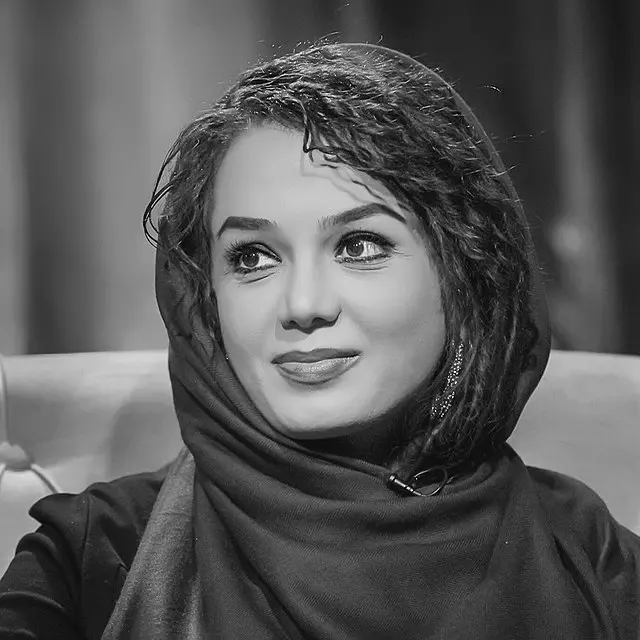 A black and white photo of a woman wearing a headscarf, smiling softly and looking to the side while seated. The background is blurred, drawing focus to her face.