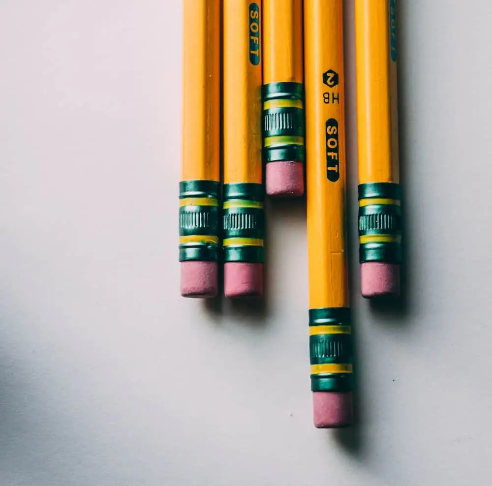 Five yellow pencils with pink erasers and green metal bands are arranged side by side on a white surface, with the erasers facing downward.